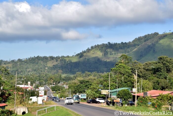 view of a small town with cars driving on a road and mountains in the background