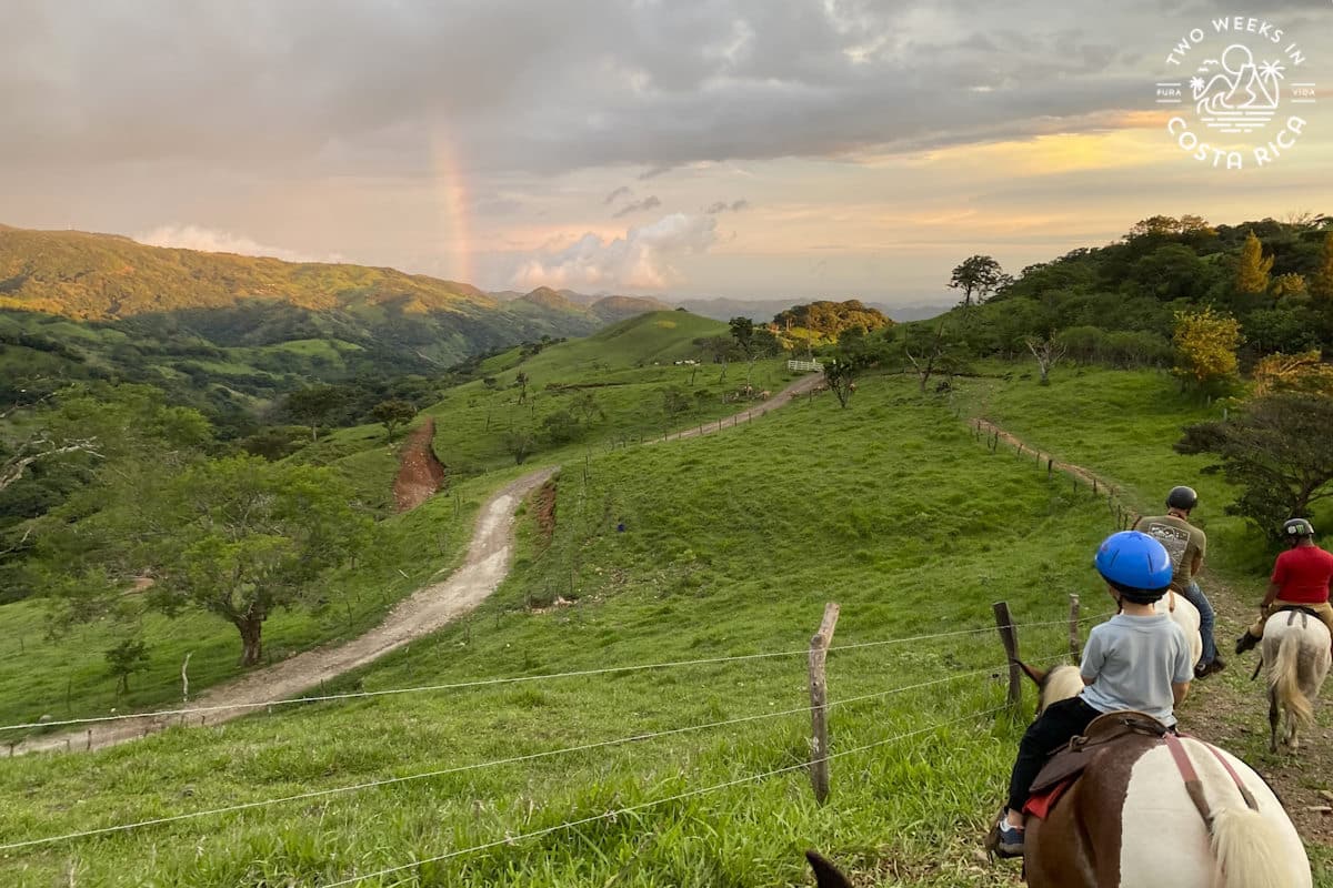 People horseback riding on a mountain ridge with a rainbow in the distance