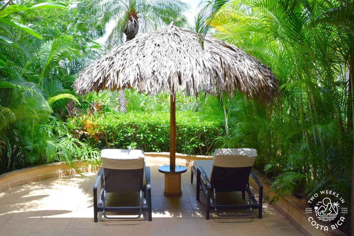 two lounge chairs under a palapa umbrella facing gardens